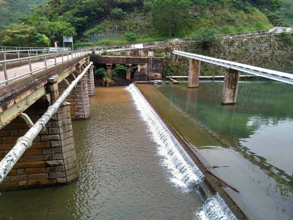 Water spilling down a stepped channel beside an old stone bridge, with pipes and wooded slopes.