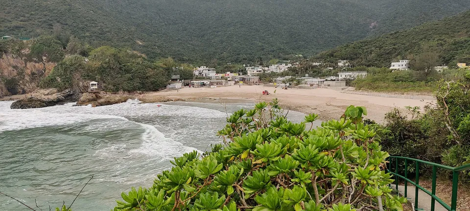 A small sandy bay with waves, shrubs in the foreground and village houses below wooded hills.