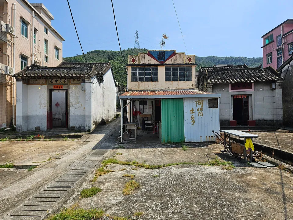 Three old Chinese-style village houses around a concrete yard, with a small shed marked 河背土地 and wooded hills behind.