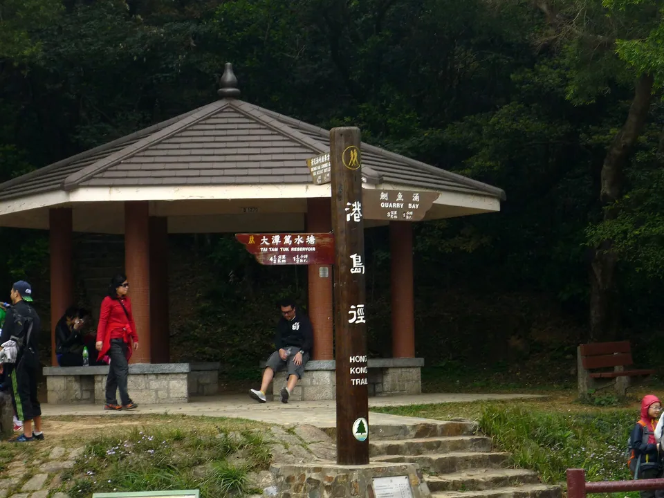 A roofed pavilion with seated hikers and a wooden fingerpost at a path junction.