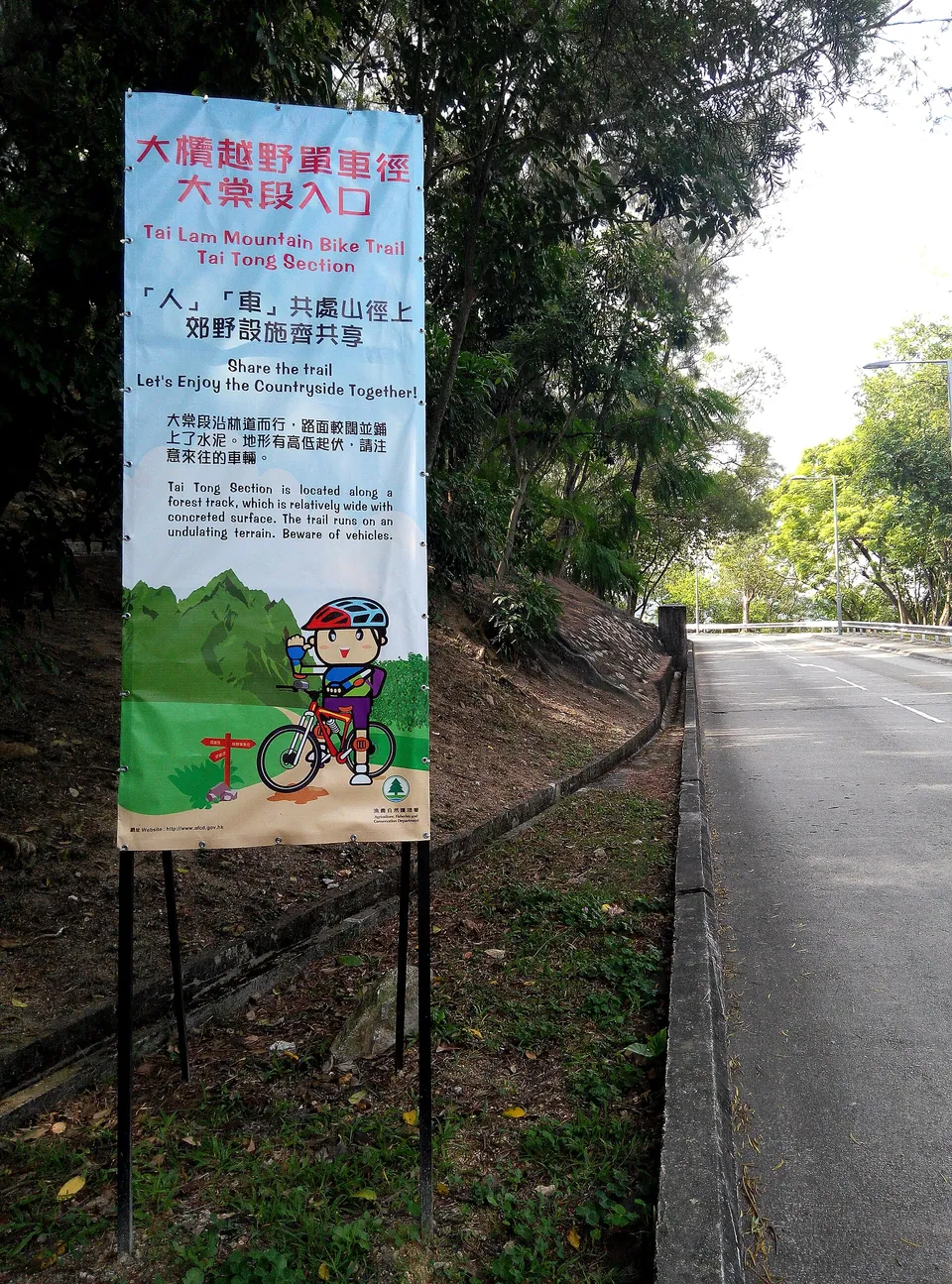 A tall illustrated signboard for the Tai Lam mountain bike trail beside a tree-lined road.