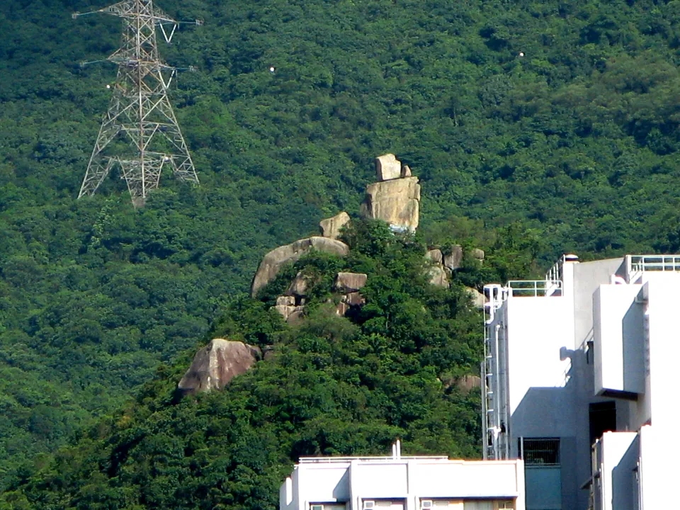 A tall rock formation on a wooded hillside above white buildings, with a pylon behind.