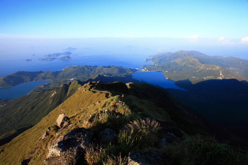 View from a high grassy ridge over a reservoir, a coastal bay and distant islands under a blue sky.