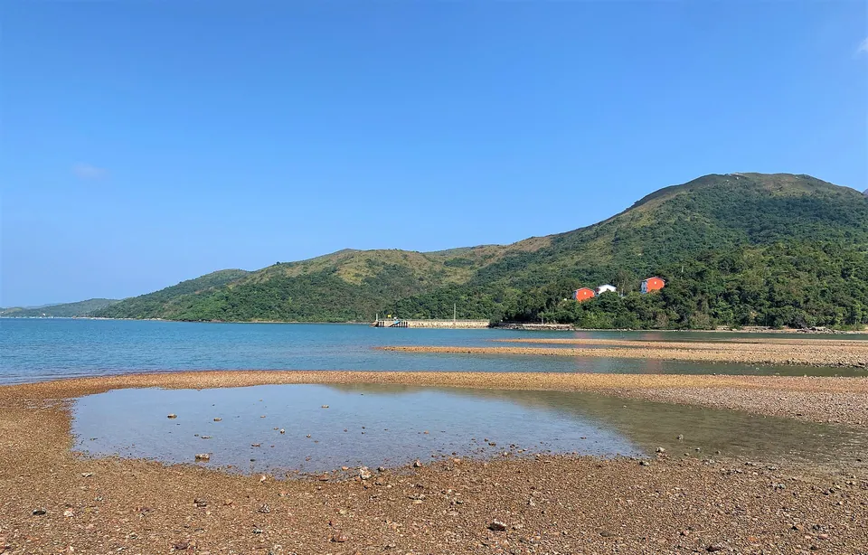 A shingle beach and shallow water in front of a green hill under a blue sky.